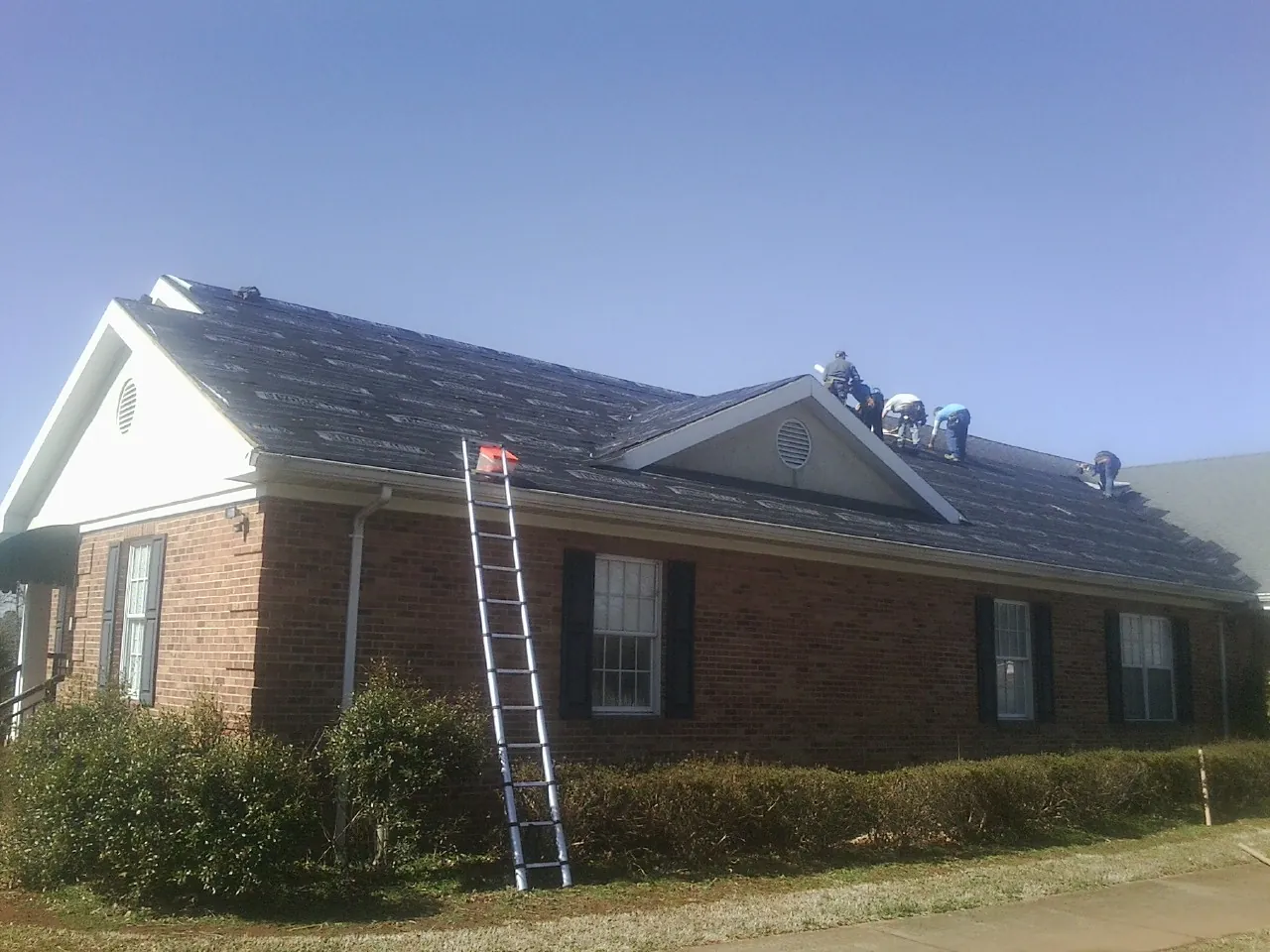Crew tearing shingles off a brick ranch roof with a ladder set — Metal Roof Repair work by our crew in Alma